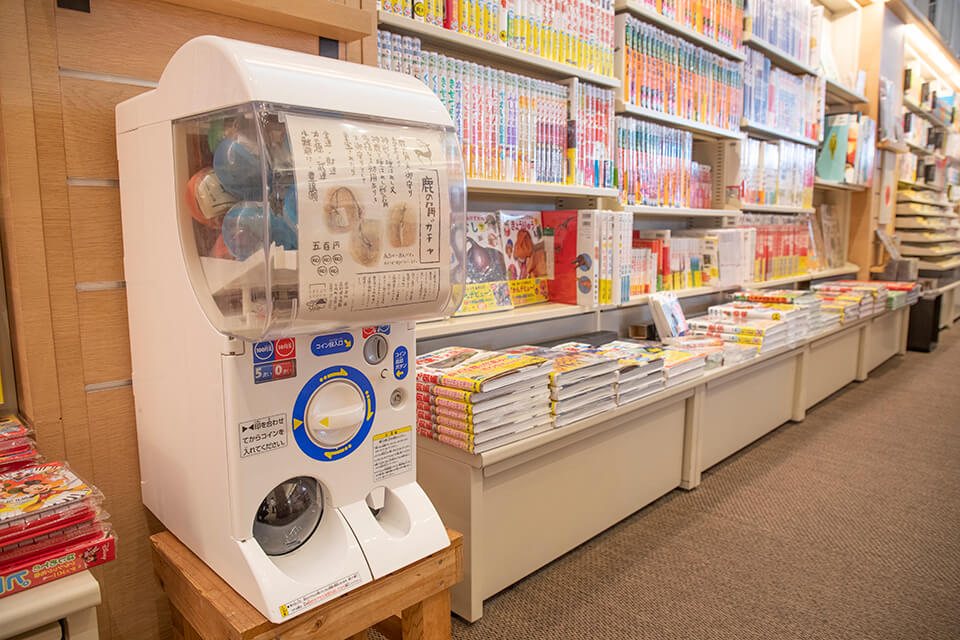 高知蔦屋書店3階の子どもの本売り場に置かれた鹿の角ガチャ。絵本や児童書の棚の横に、白いガチャが立っている。
